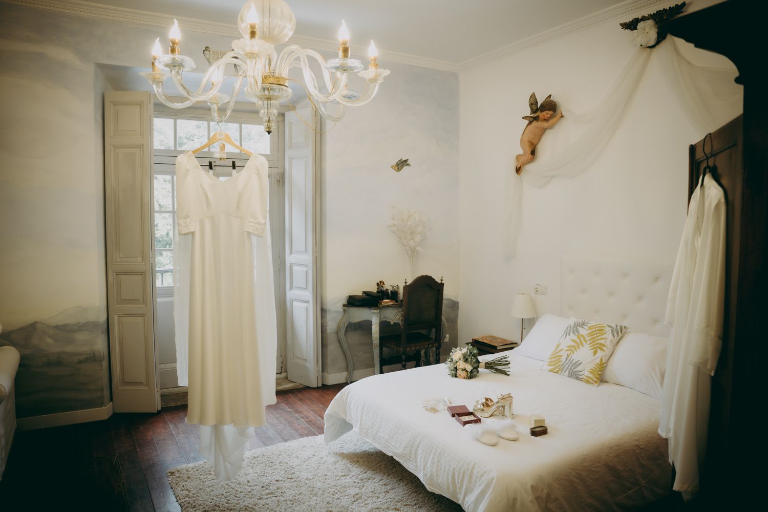Elegante sala de preparación nupcial con vestido de novia blanco, cama con dosel y lámpara de araña de cristal con luz natural.