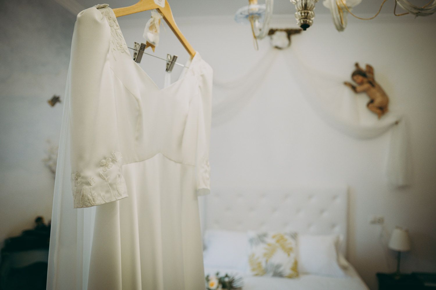 Un elegante vestido de novia blanco cuelga de una percha en un dormitorio suavemente iluminado con ropa de cama floral.