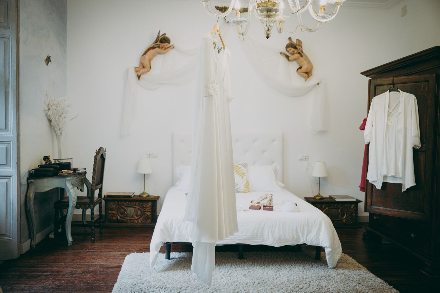 Elegante dormitorio blanco con decoración vintage que incluye apliques de pared dorados ornamentados, cama con dosel y muebles antiguos.