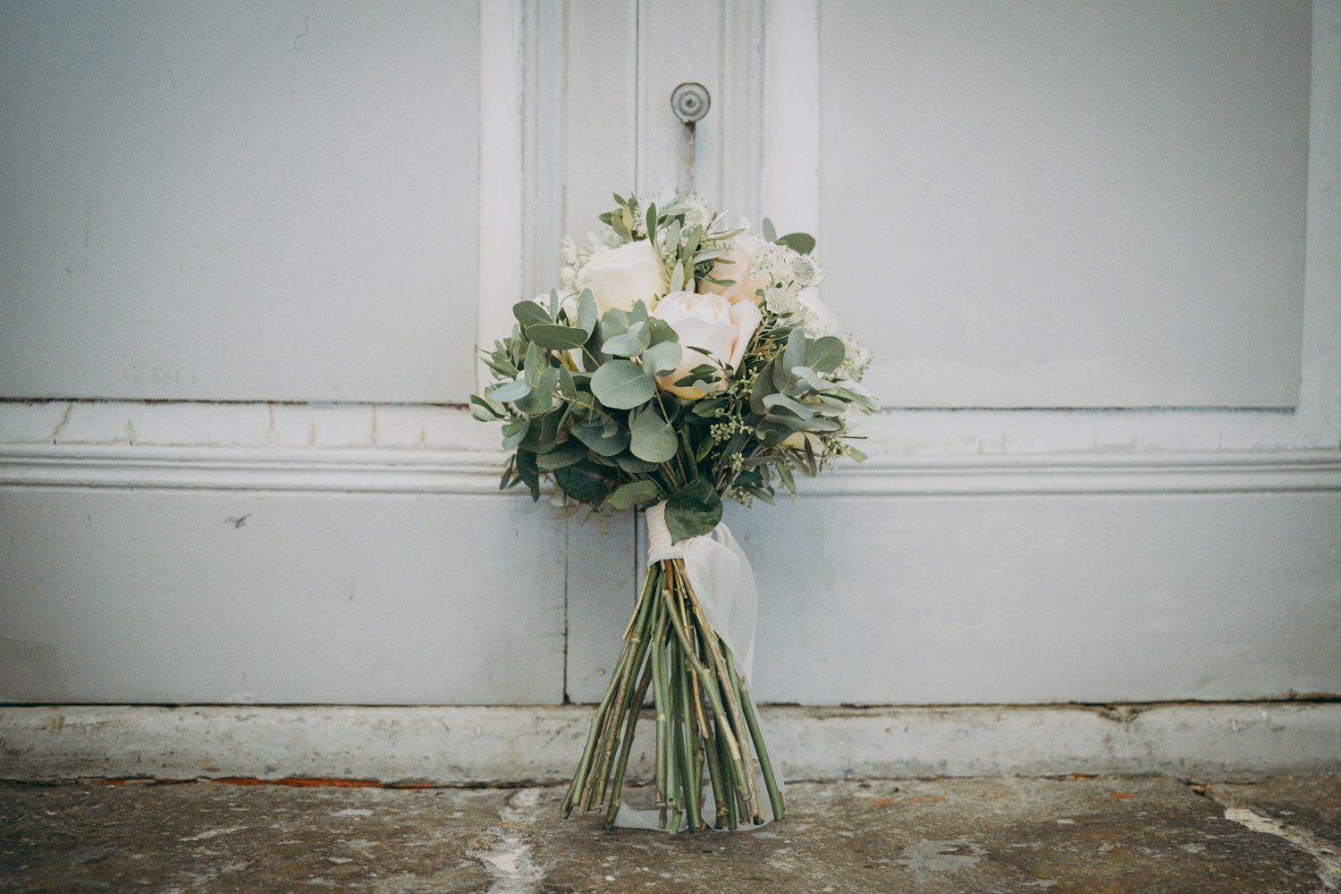 Un delicado ramo de novia con rosas blancas y hojas de eucalipto destaca sobre una pared gris claro.