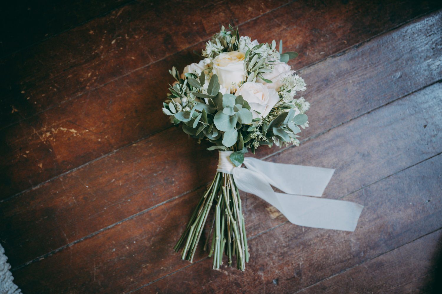 Un elegante ramo de novia con rosas blancas y eucalipto atado con una cinta blanca descansa sobre un suelo de madera oscura.