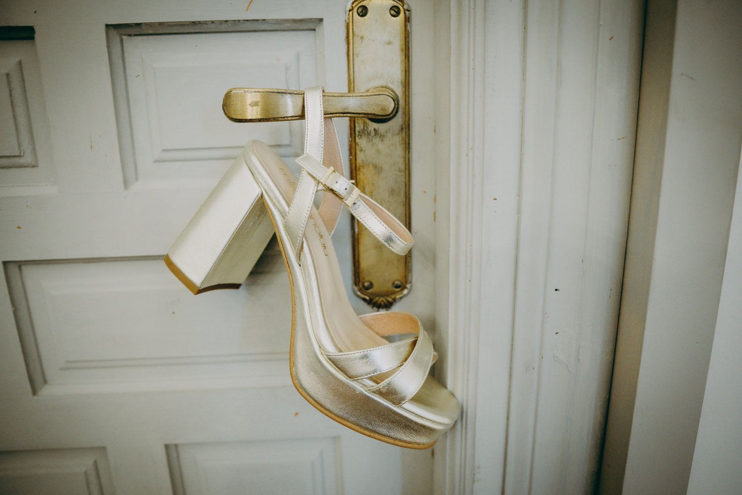 Un par de elegantes sandalias de novia blancas de tacón alto colgadas de una manija de puerta vintage.