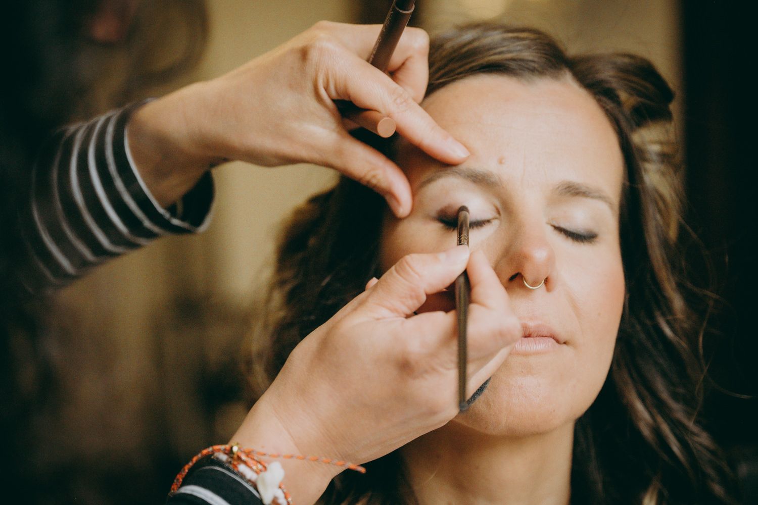 Una maquilladora con mangas a rayas aplica cosméticos a una modelo con los ojos cerrados bajo una iluminación cálida.
