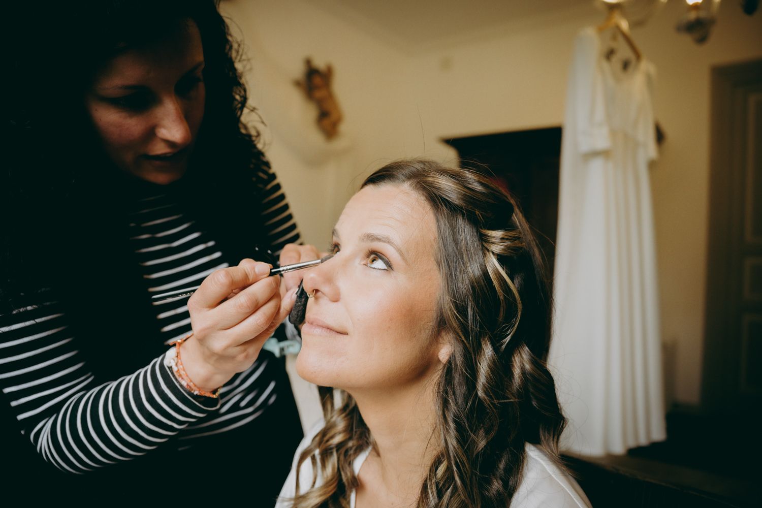 Maquilladora profesional aplicando cosméticos con un vestido de novia blanco visible en el fondo.