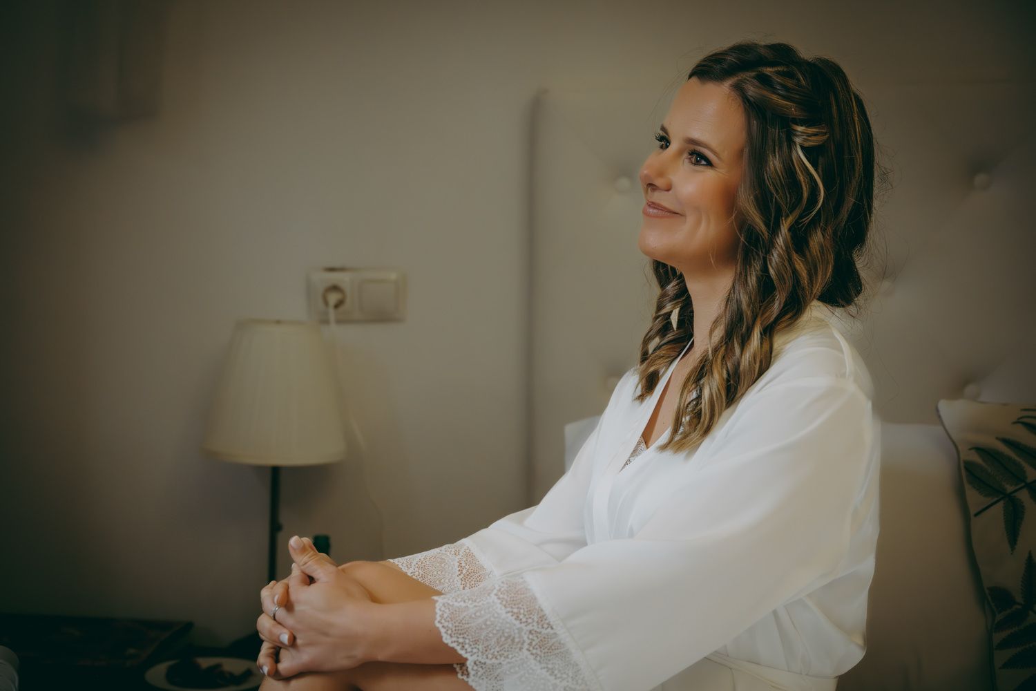 Una novia vestida con una túnica blanca sonríe mientras está sentada en una habitación con una lámpara al fondo.
