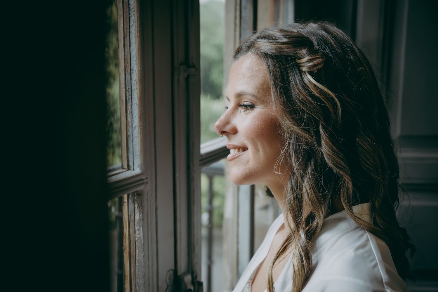 Una novia vestida de blanco mira hacia fuera por una ventana por la que entra luz natural.