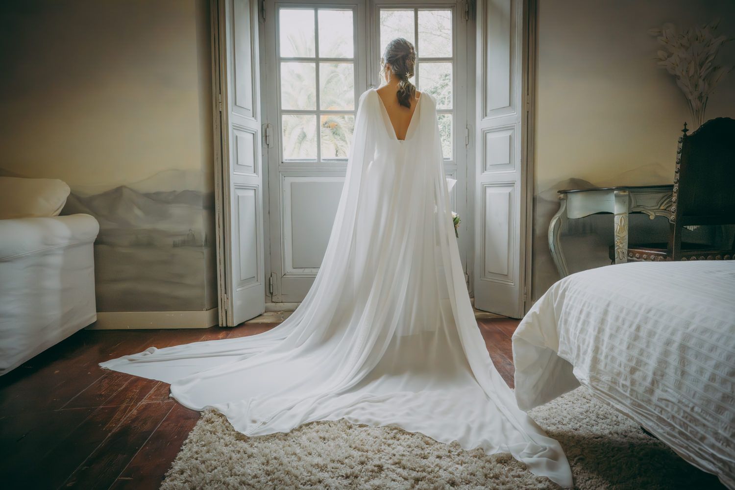 Elegante silueta de novia en vestido blanco fluido frente a puertas francesas y luz natural en el palacio de Villabona. Asturias.