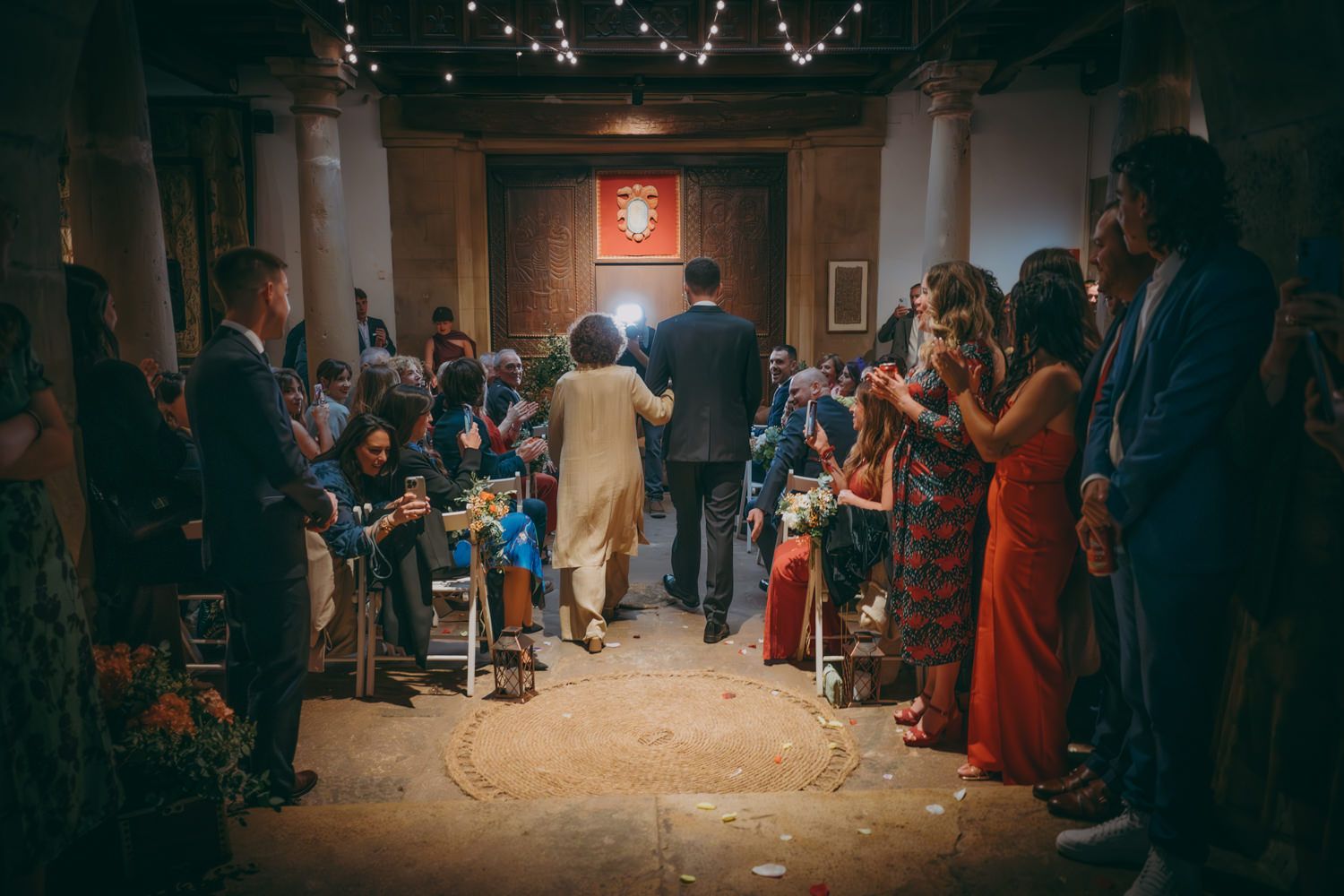 Ceremonia de boda en un lugar rústico con luces de cadena en el techo mientras los invitados se alinean en el pasillo.