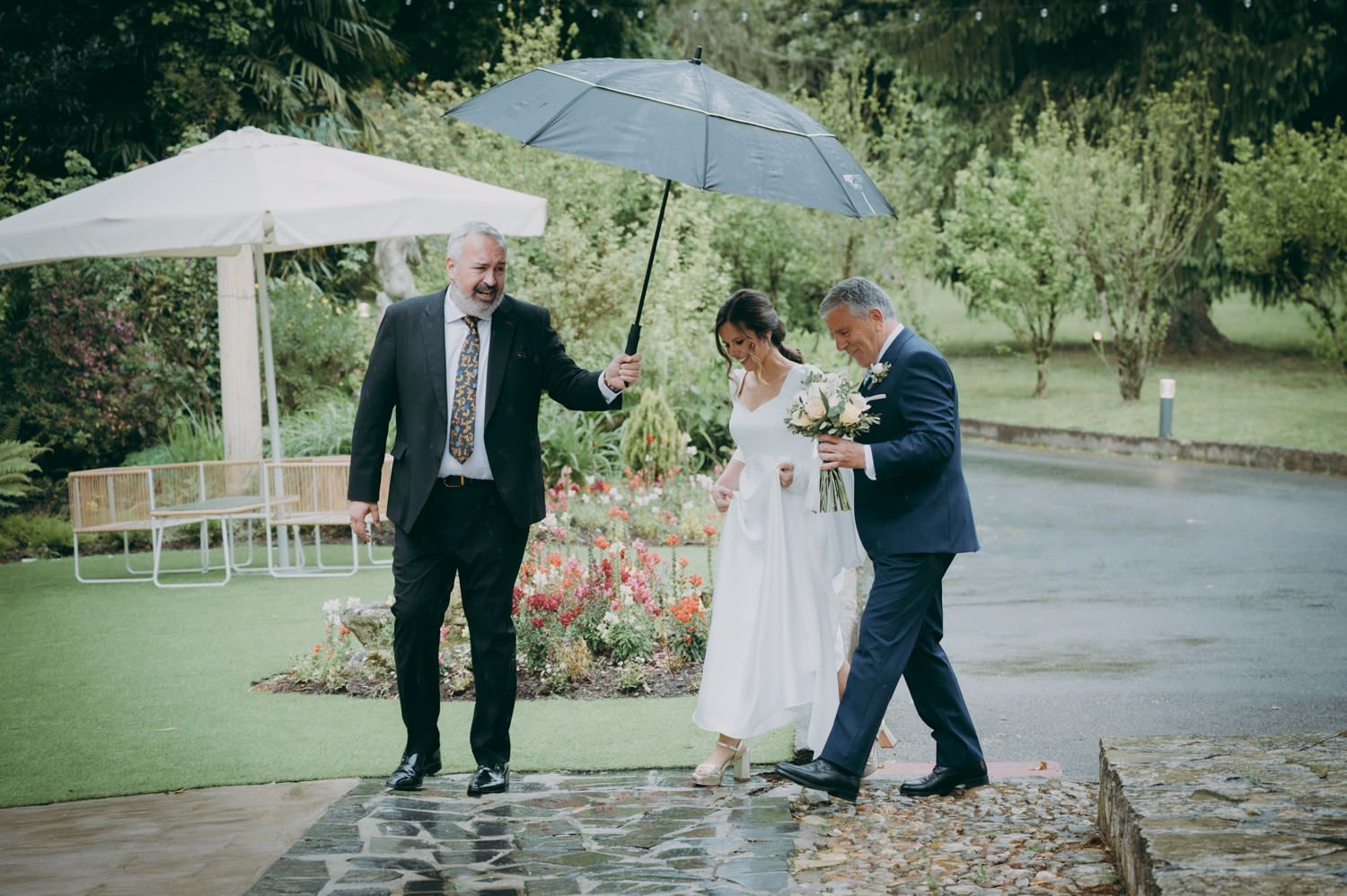 Fiesta de bodas caminando bajo paraguas en un jardín en un día lluvioso.