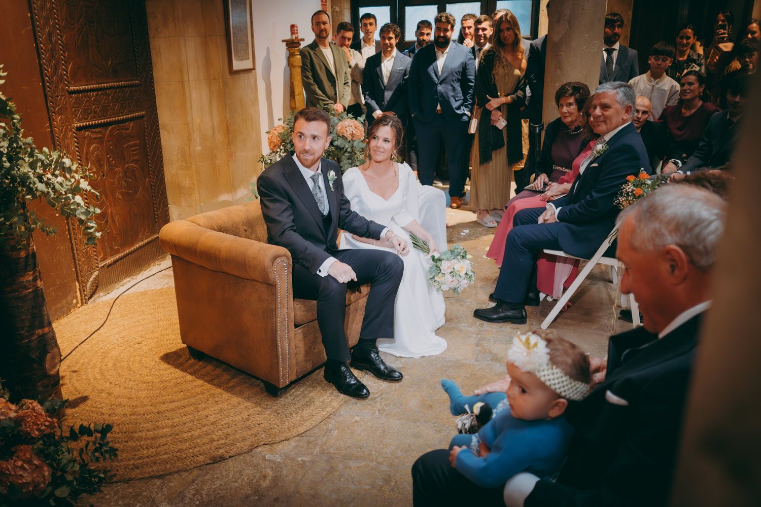 Recién casados sentados en una silla de cuero durante una ceremonia íntima con invitados mirando.