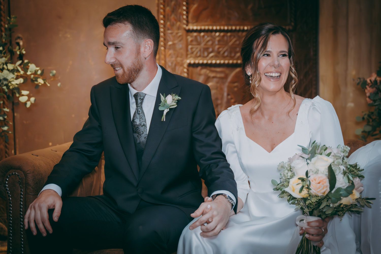 Pareja compartiendo un momento tierno en un banco de cuero adornado durante la ceremonia de la boda.