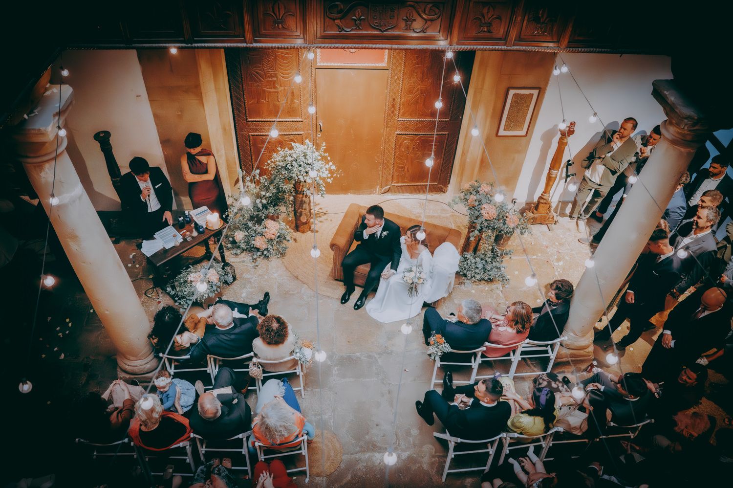 Fotografía cenital de una ceremonia nupcial con invitados sentados en sillas alrededor de columnas ornamentadas.