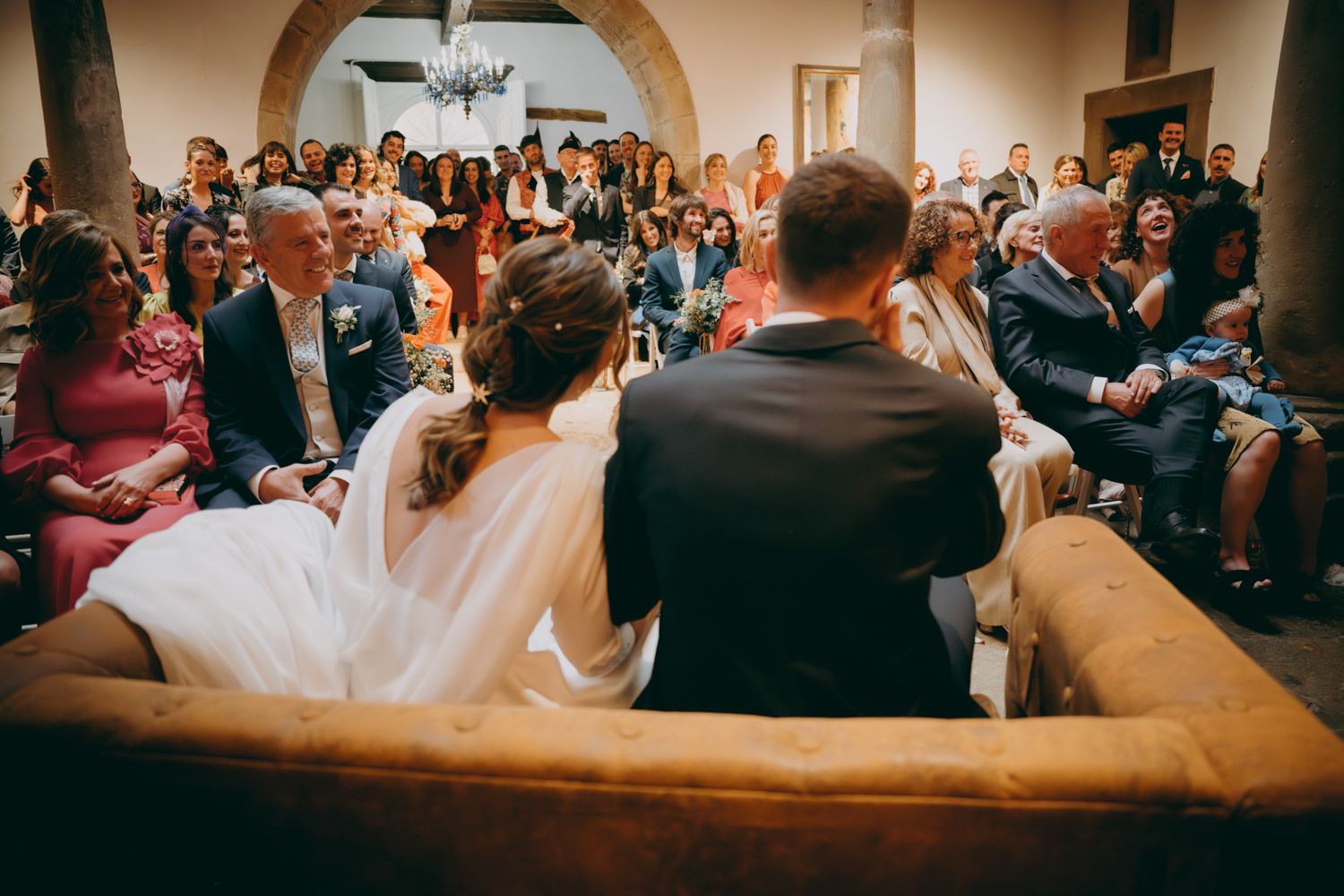 Los invitados a la boda llenan filas de asientos en un lugar interior ornamentado con arquitectura arqueada.