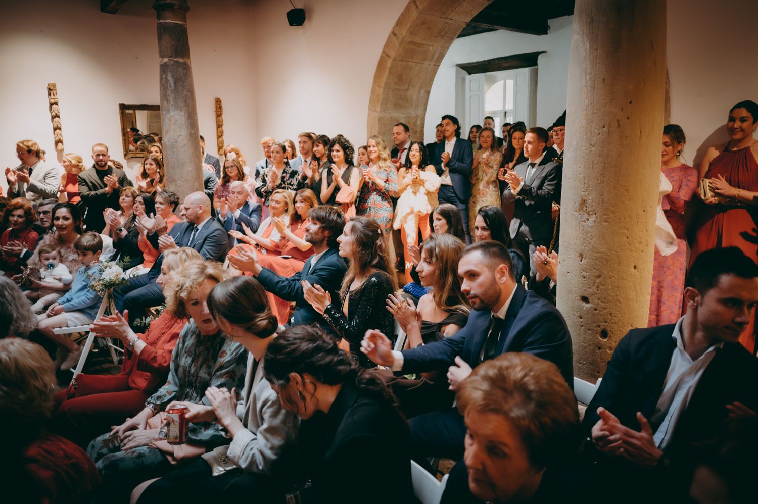 Multitud de invitados a la boda sentados en un lugar histórico con arcos de piedra.