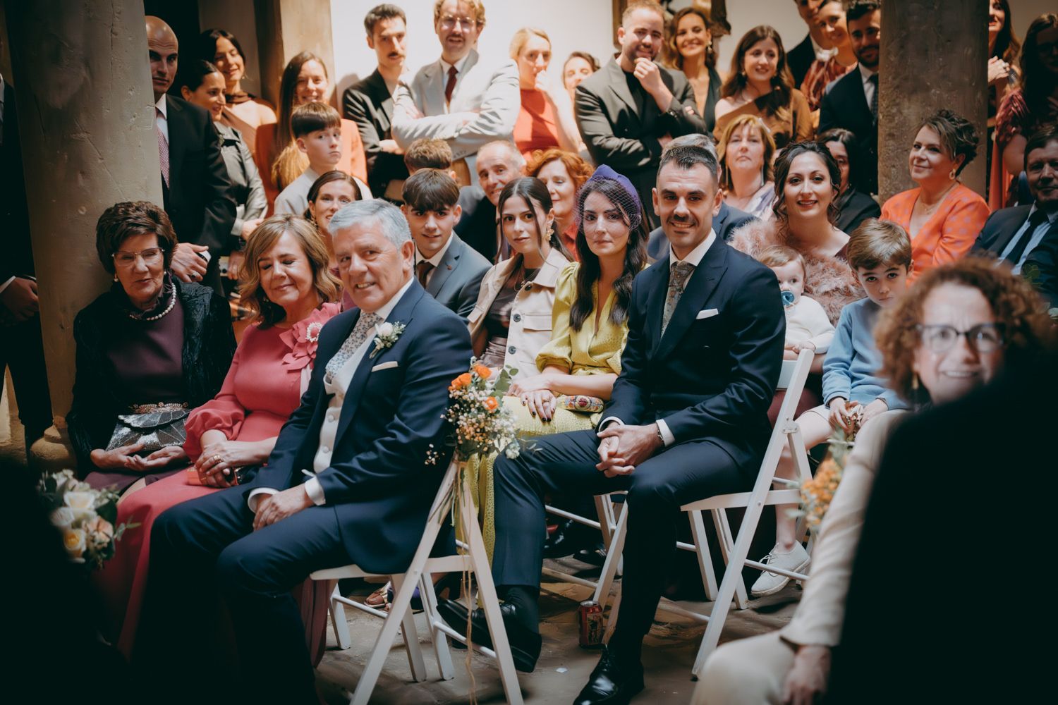 Los invitados a la boda llenan filas de sillas blancas durante una ceremonia en el interior.