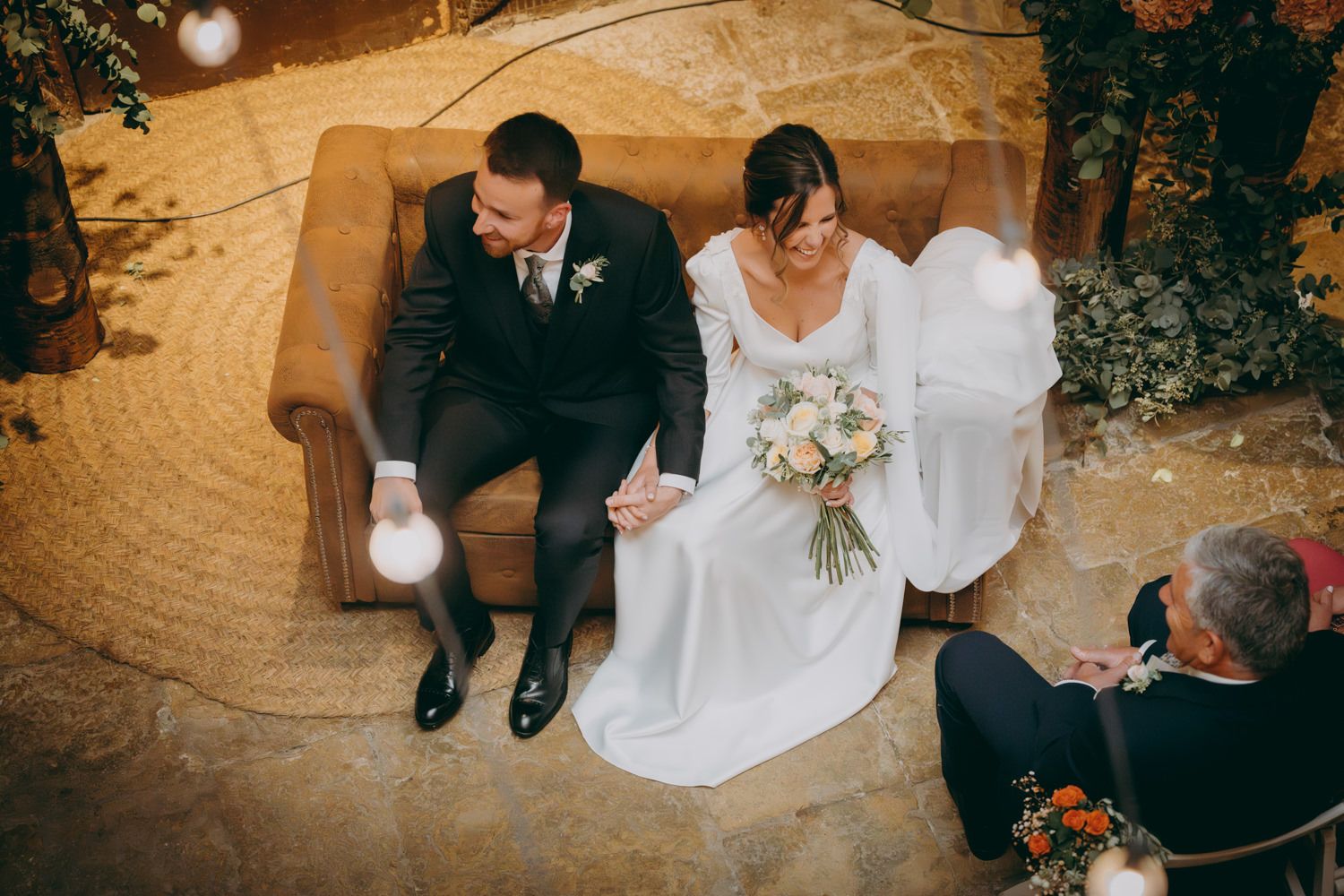 Recién casados sentados en un sofá marrón adornado durante su ceremonia de boda.