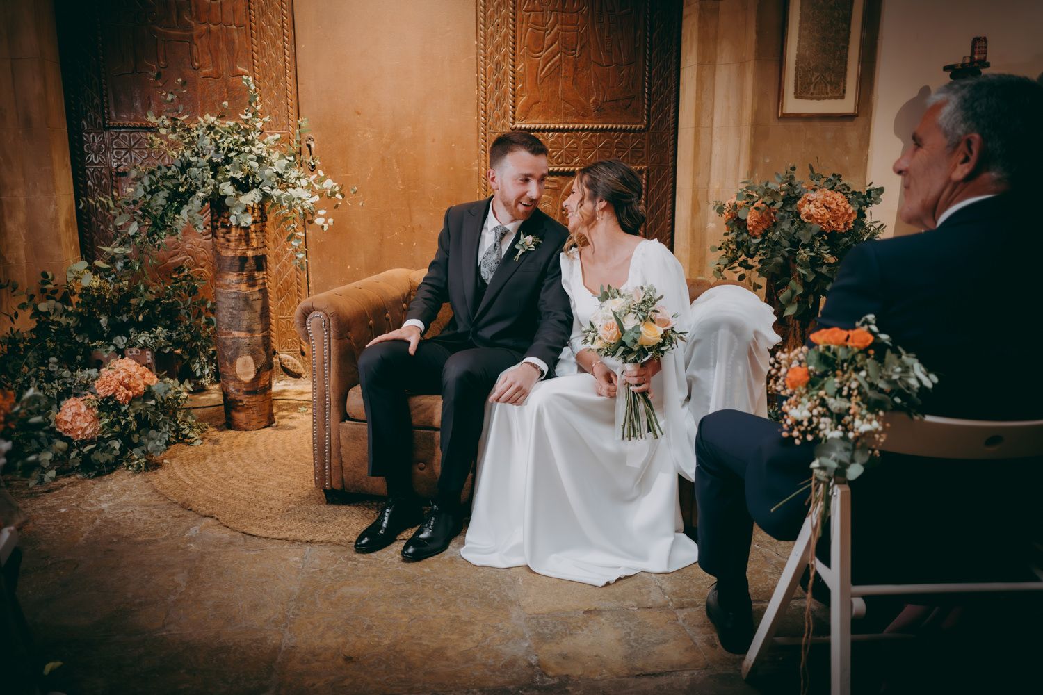 Una pareja de novios comparte un momento íntimo en un sofá de cuero rodeado de arreglos florales.