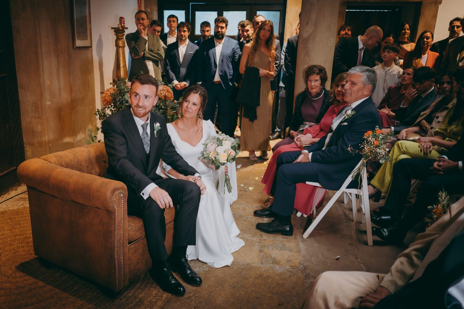 Novia y novio sentados en un sofá de cuero marrón durante una ceremonia en el interior con invitados.