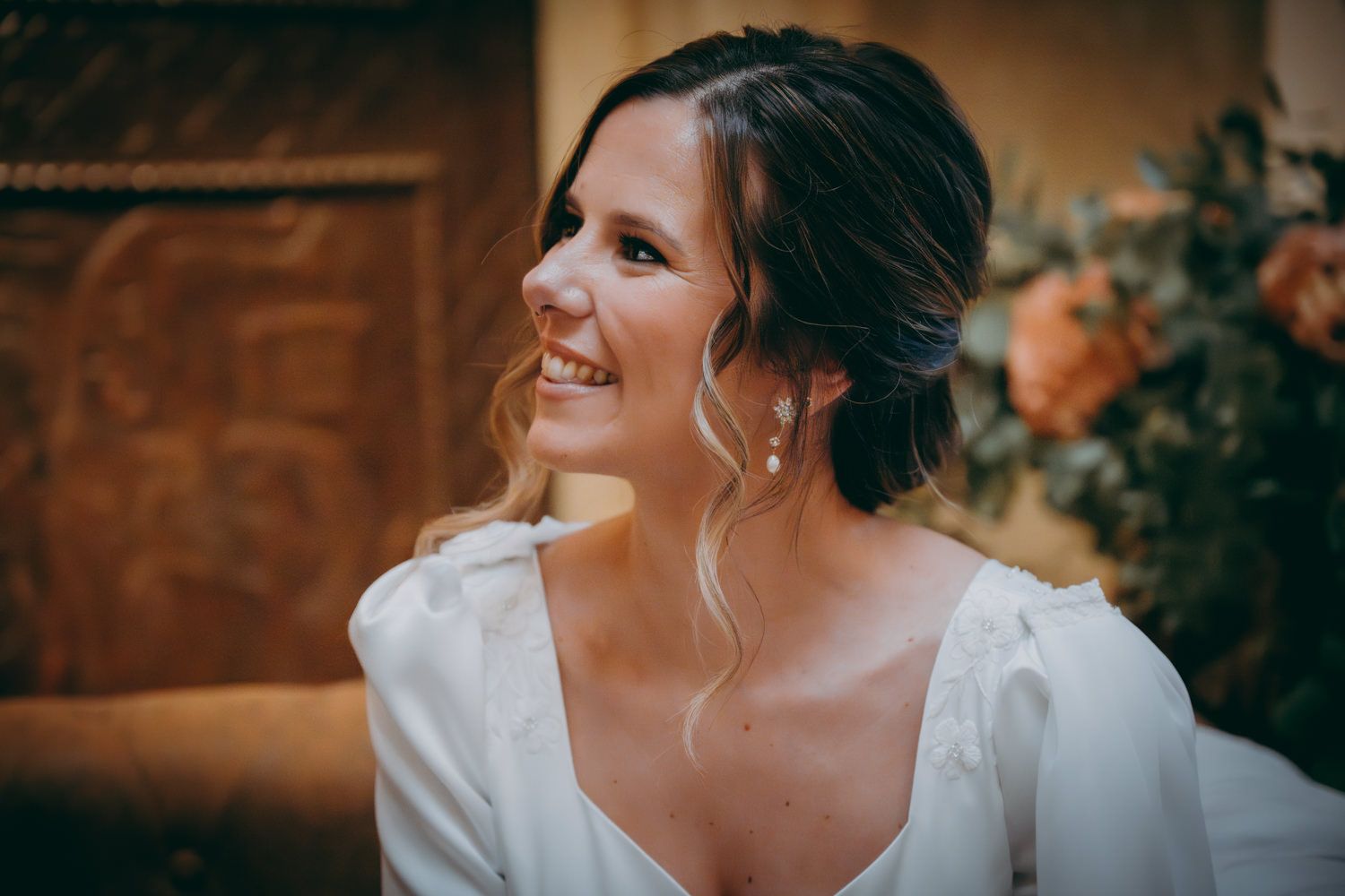 Perfil lateral de una novia con un vestido blanco clásico sonriendo cálidamente con un elegante peinado recogido y pendientes colgantes.