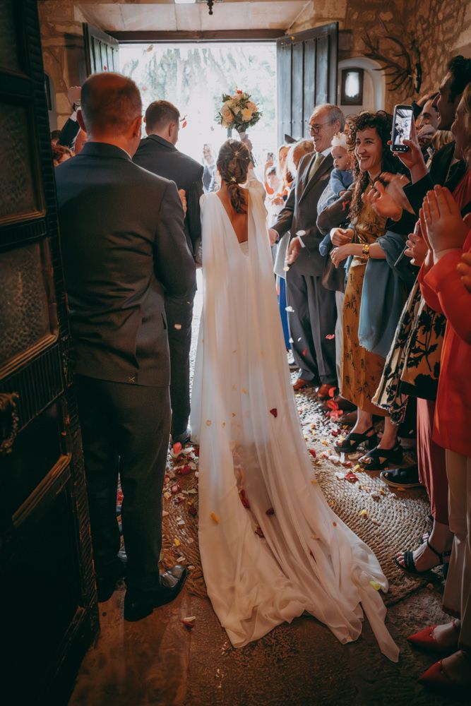 Una pareja de novios camina por un pasillo lleno de invitados mientras cae confeti en un lugar de piedra.