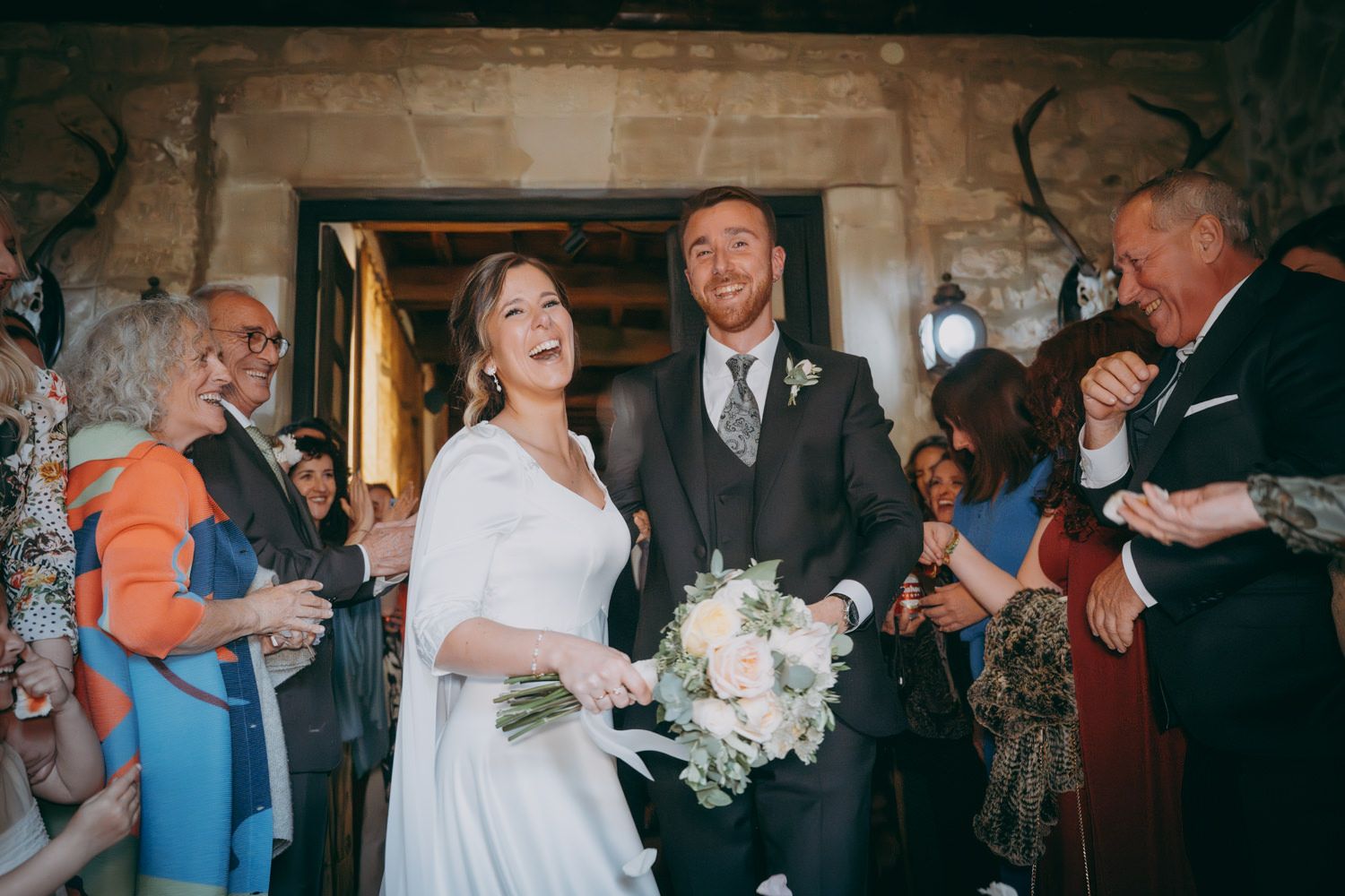 Una pareja celebra el día de su boda en un lugar con paredes de piedra y rodeado de invitados.