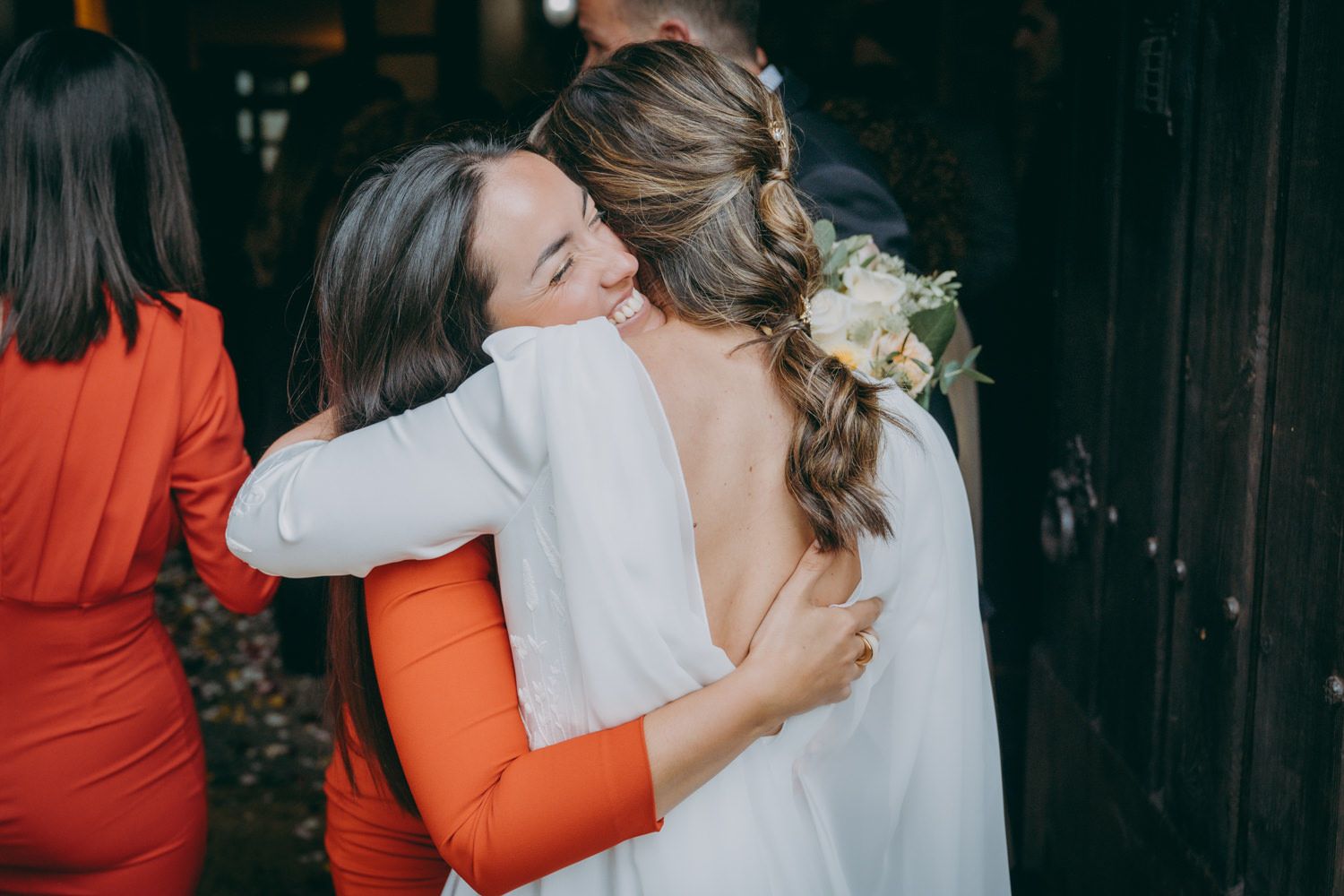 Dos personas comparten un cálido abrazo en una boda, una vestida de naranja y otra de blanco.