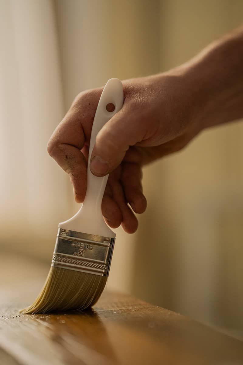 Close-up of a hand holding a paintbrush while staining or varnishing wood.