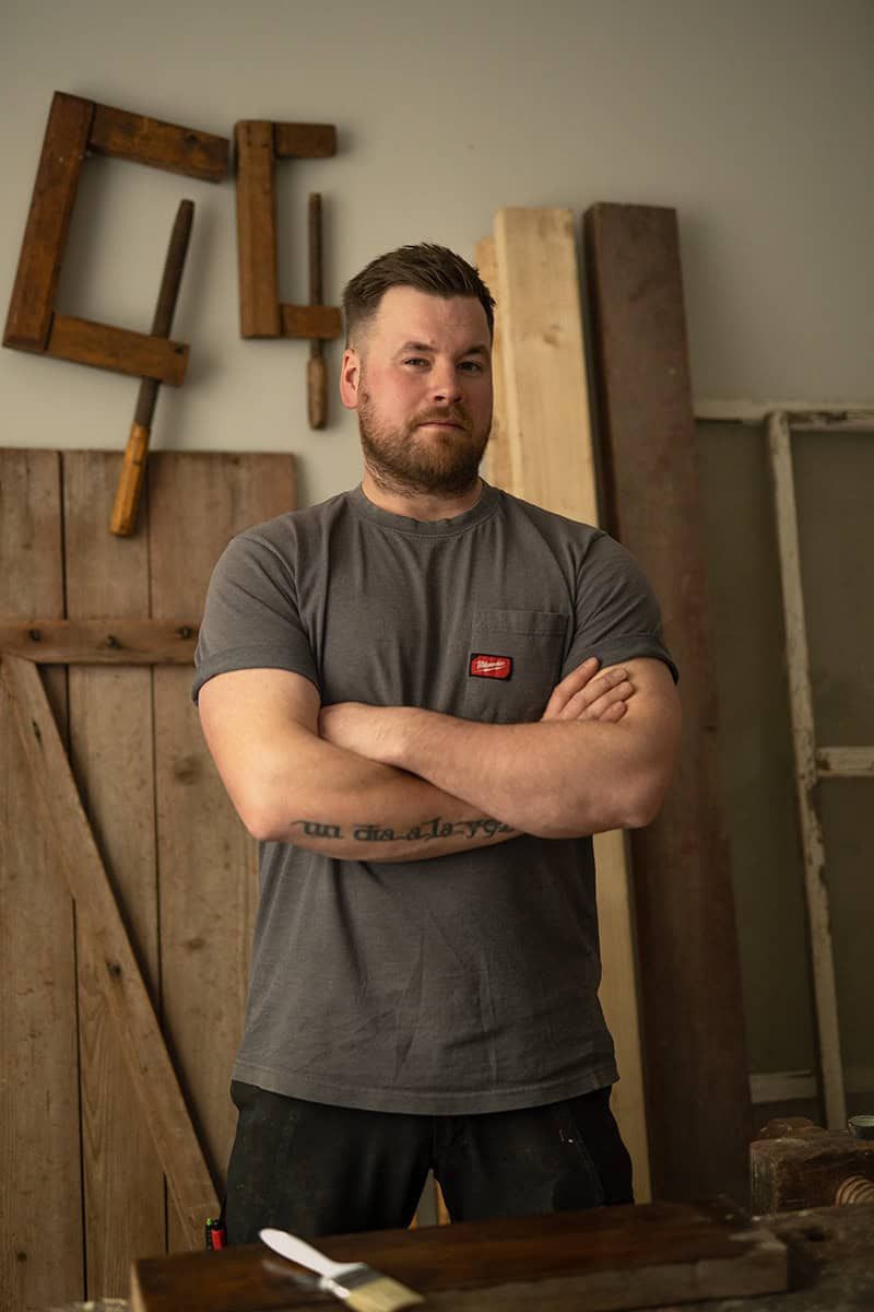 A craftsman in a gray t-shirt standing in front of wooden tools and materials.