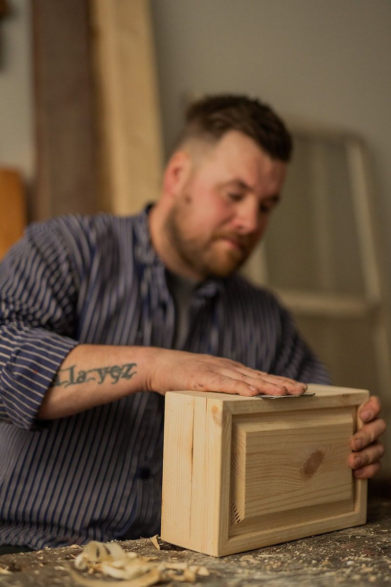 Craftsman carefully examining a wooden box on a workbench covered in wood shavings.