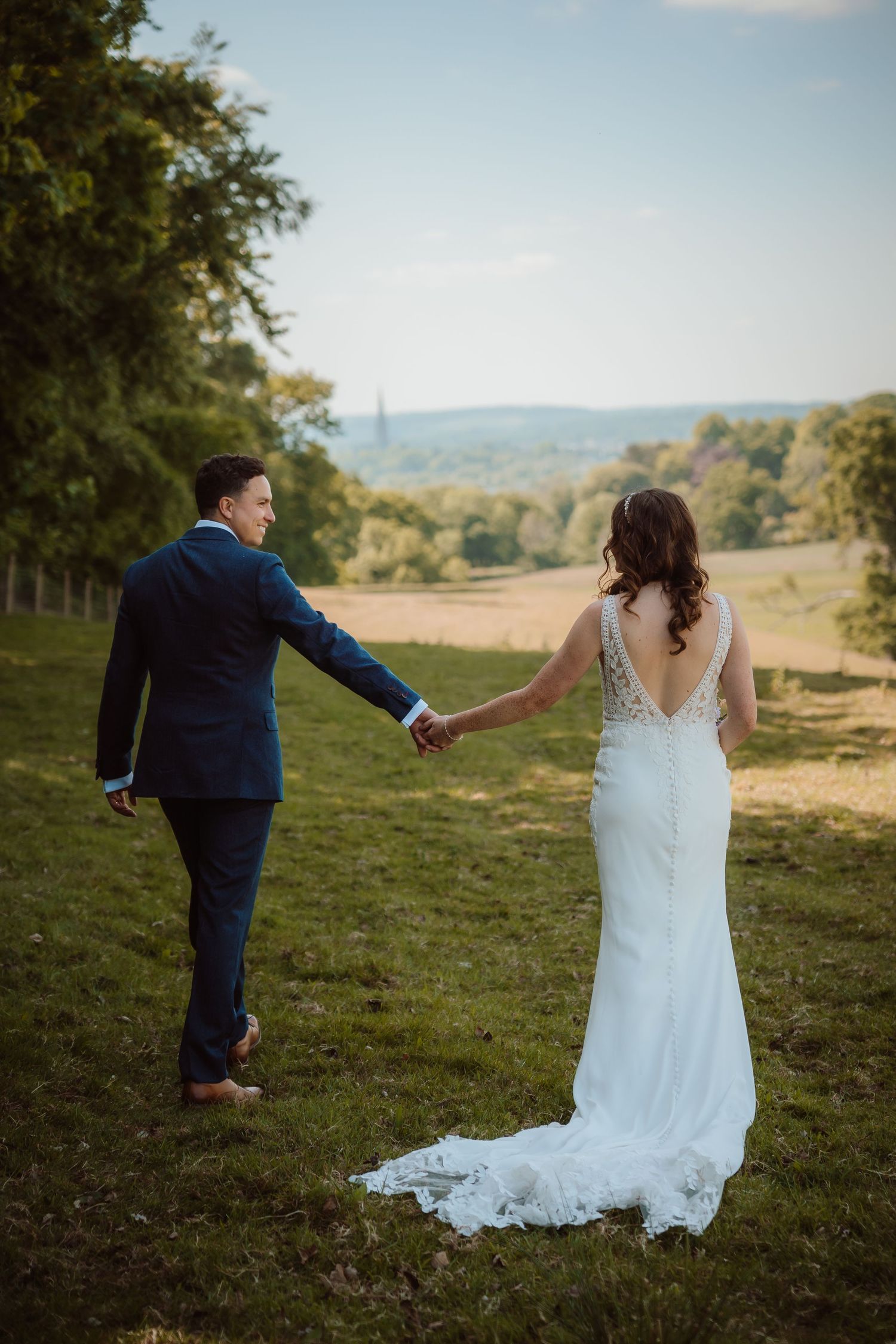 A couple in wedding attire holds hands while walking up a grassy hill with a scenic countryside view in the background.
