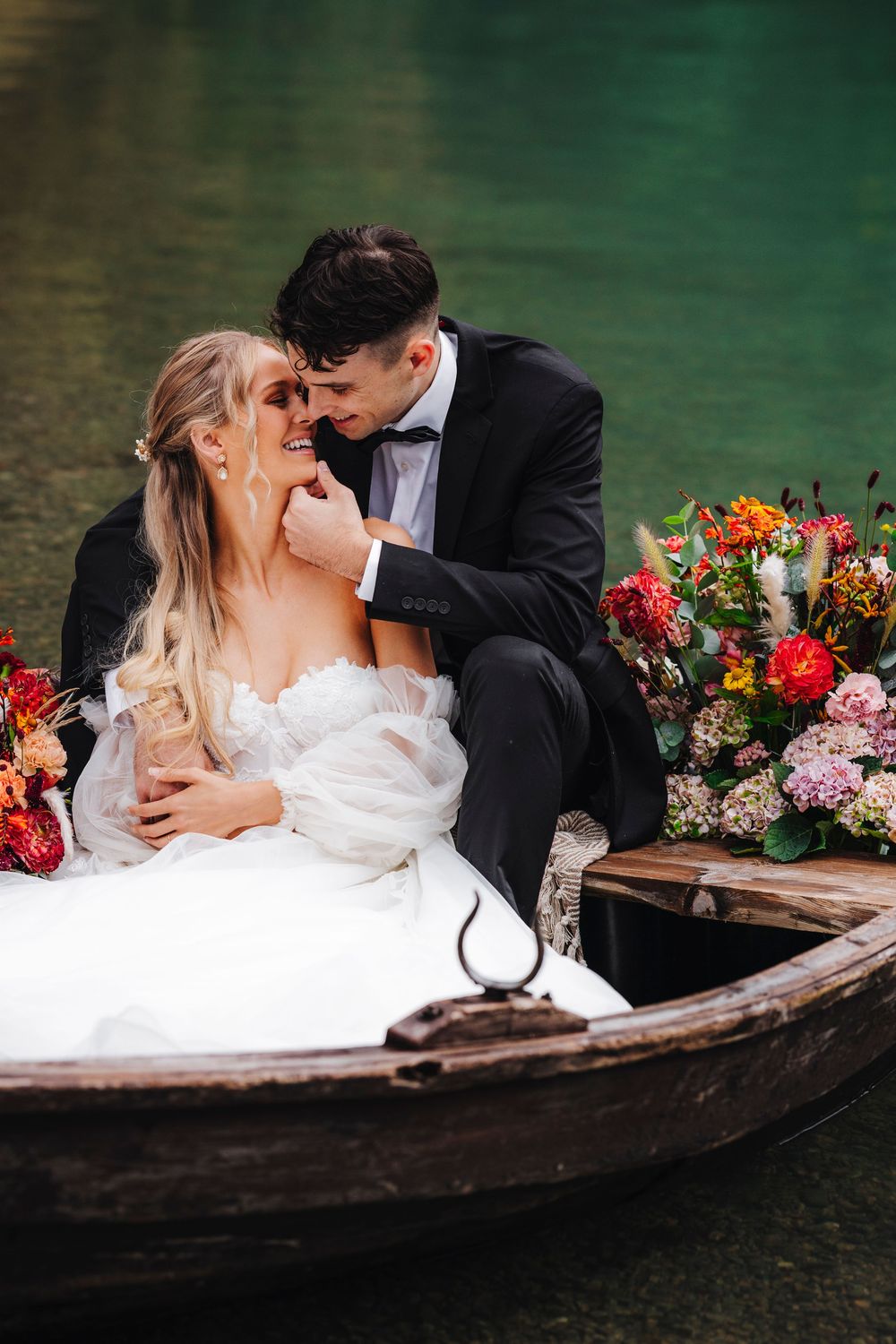 Romantic wedding couple sharing intimate moment in wooden boat with vibrant floral arrangement on emerald waters.