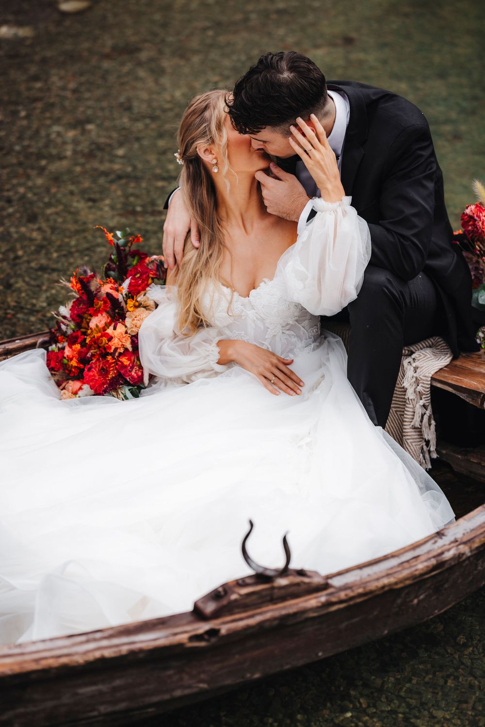 Newlywed couple shares intimate moment in rustic wooden boat with vibrant red fall floral bouquet nearby.