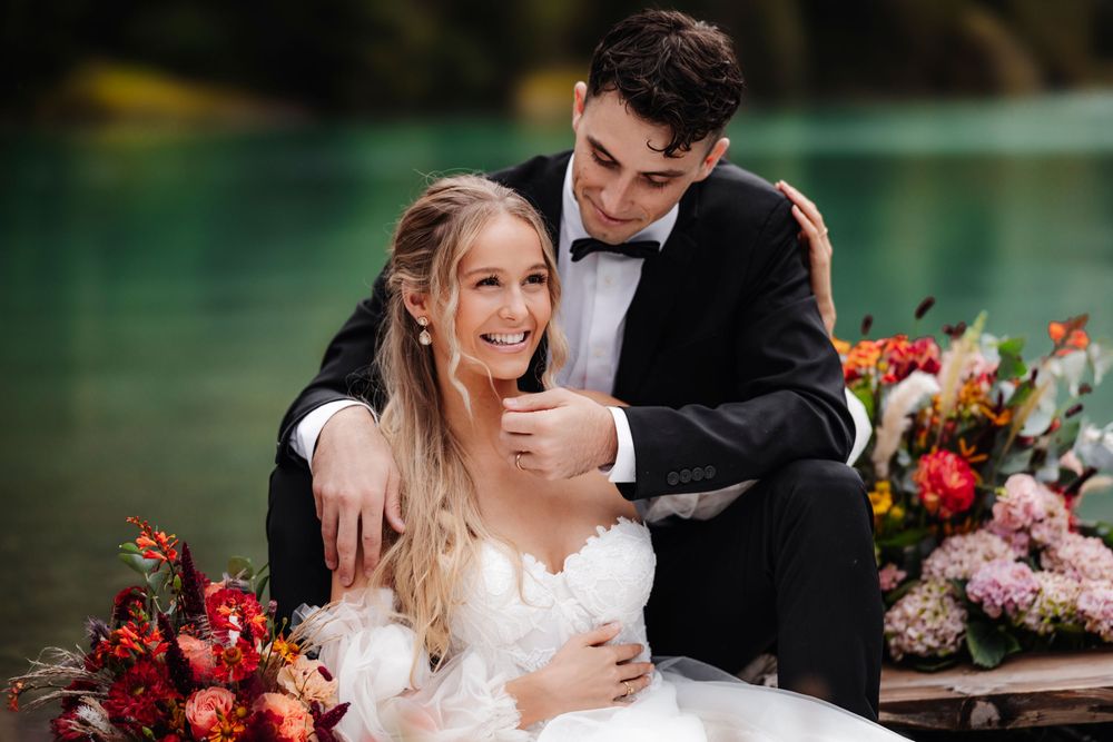 Two people in wedding attire share a joyful moment next to vibrant floral arrangements by a lake.