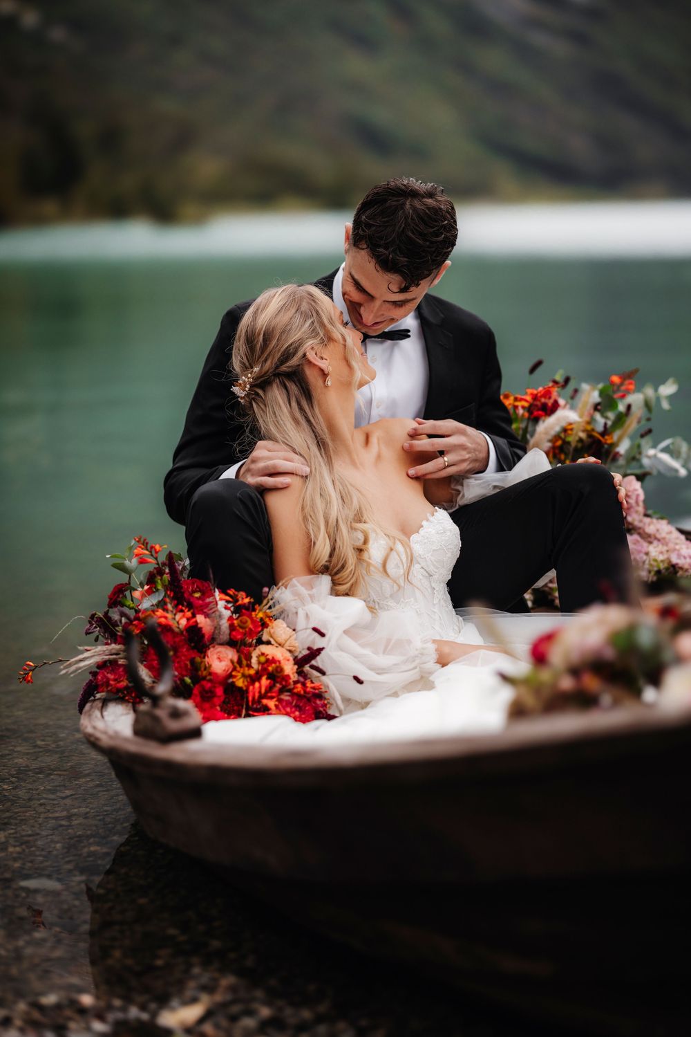 Romantic wedding couple embraces in wooden boat on tranquil mountain lake with vibrant red floral arrangements.