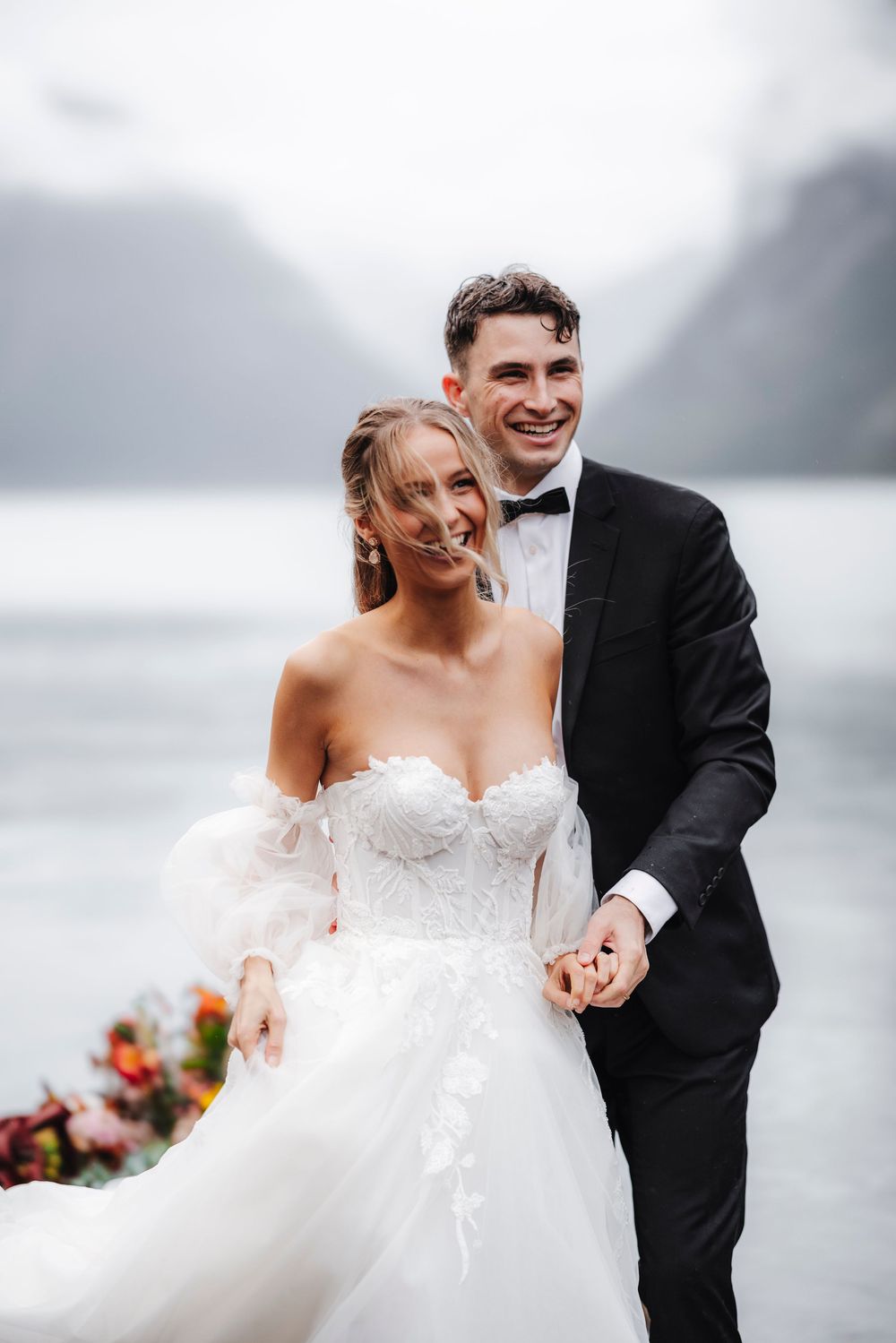 Newlyweds share a joyful moment by a misty lake, with the bride in a strapless white gown and groom in classic black tuxedo.