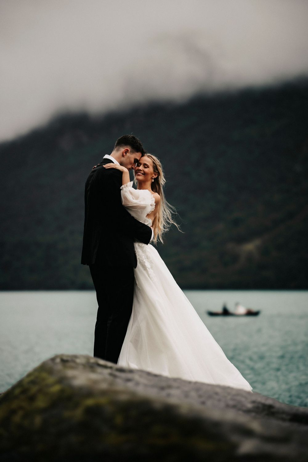 Romantic wedding couple embraces on rocky cliff with lake and mountains in moody dramatic lighting at sunset.