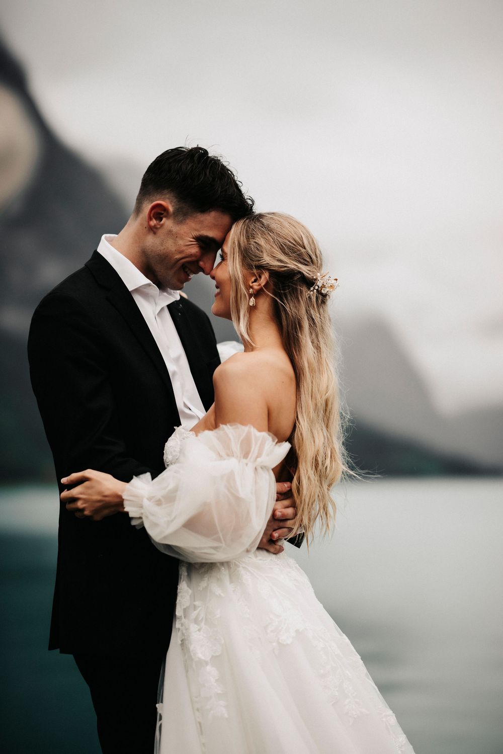 A romantic wedding photo overlooking a misty lake with mountains, showcasing a tender moment between newlyweds.