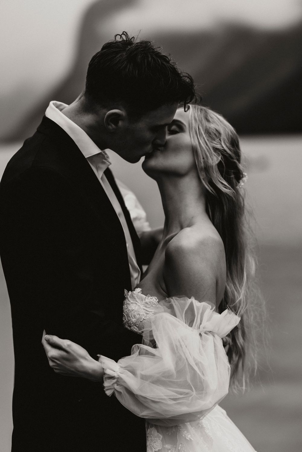 Black and white romantic wedding photo showing a couple sharing an intimate kiss against a dramatic outdoor background.