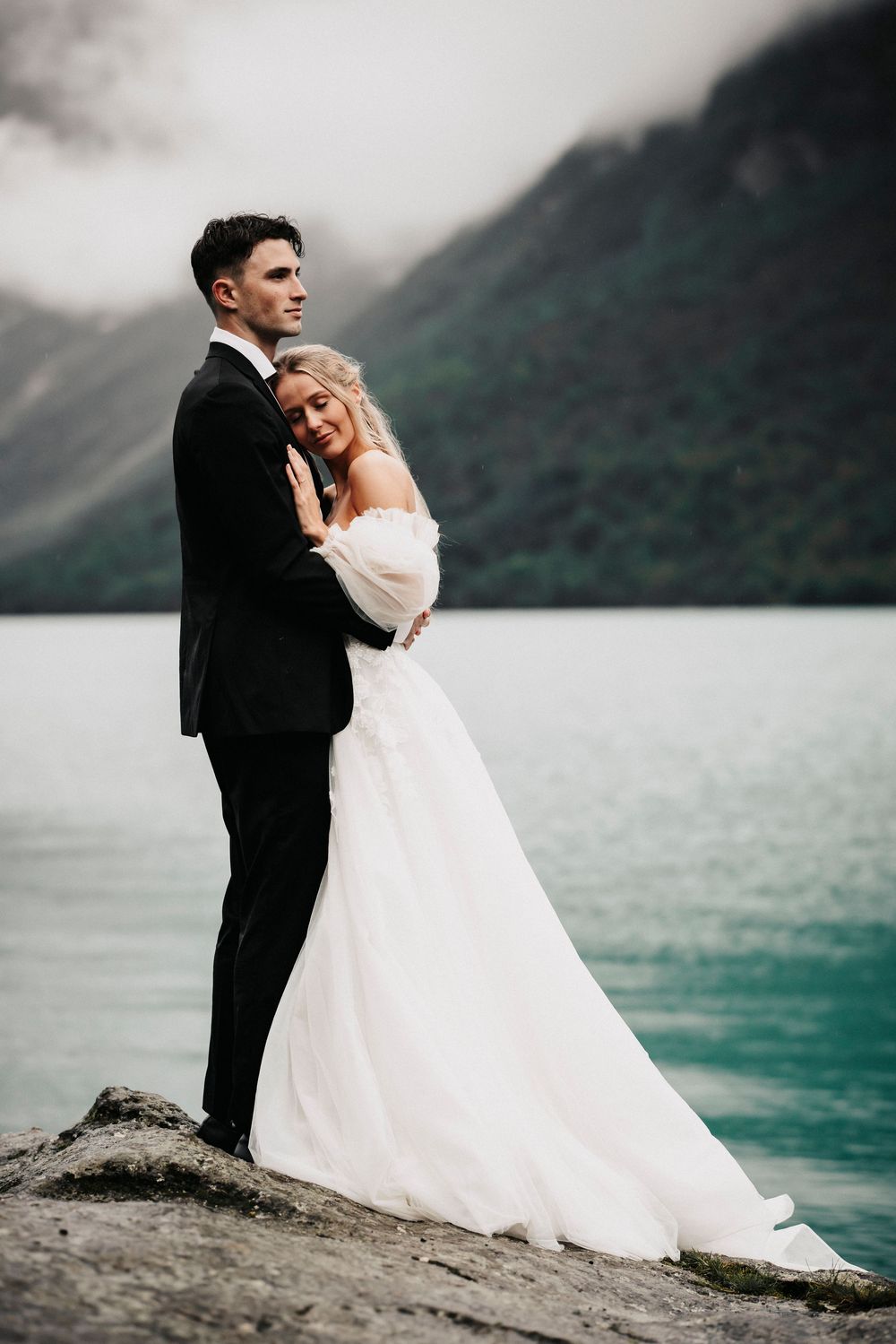 A bride and groom embrace while standing on a rocky shore with turquoise waters and misty mountains in the background.