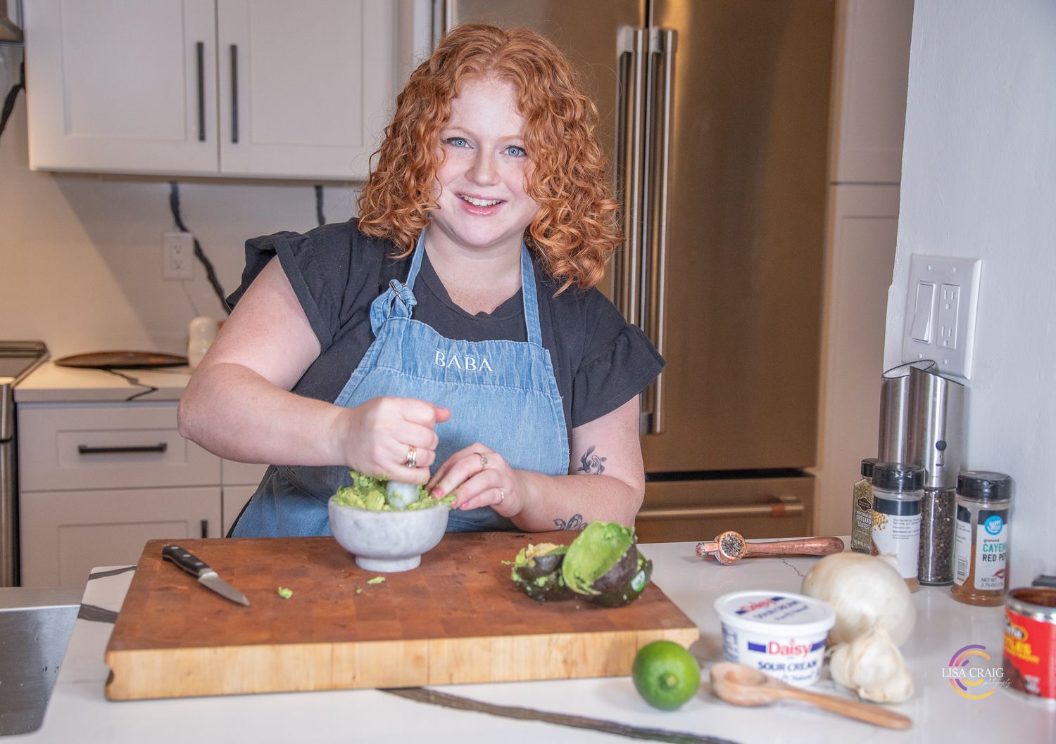 Someone in a denim apron stands at a wooden cutting board in a kitchen while preparing food with citrus and avocado.