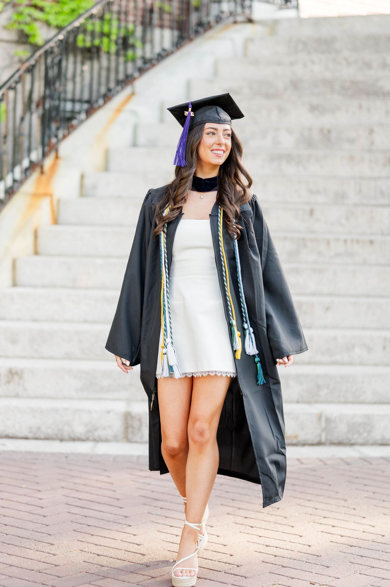 Graduate in cap and gown wearing a white dress stands on brick steps during commencement ceremony.