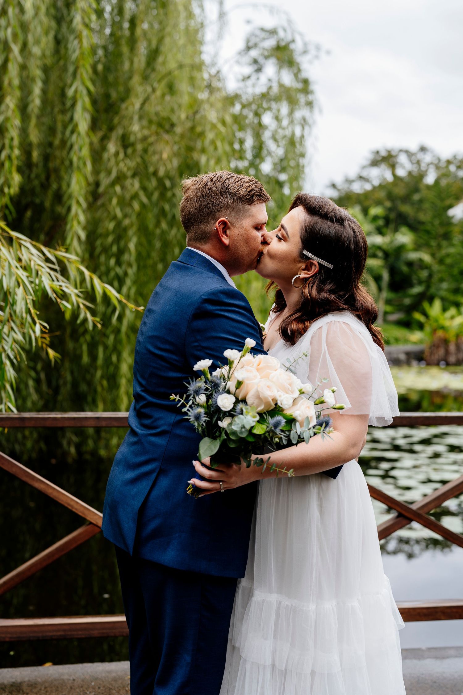 Newlyweds share a romantic kiss on wooden bridge by willow tree with white roses bouquet and elegant wedding attire.