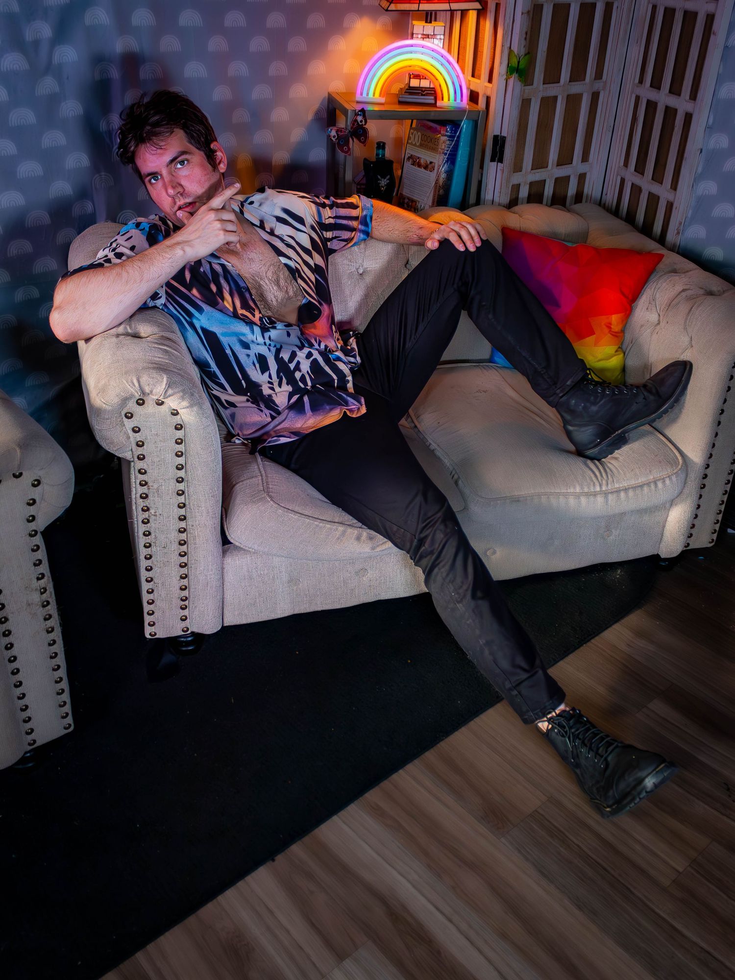 Person lounging on a gray studded couch with rainbow neon lighting and wearing a patterned shirt.