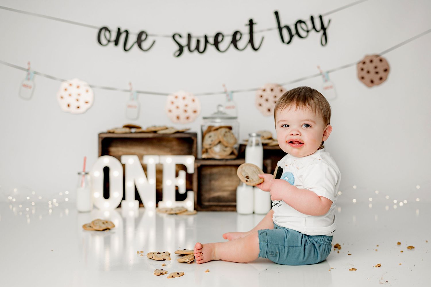 First birthday photo setup with white letters spelling ONE, rustic wooden crate, and cookie-themed party decorations.