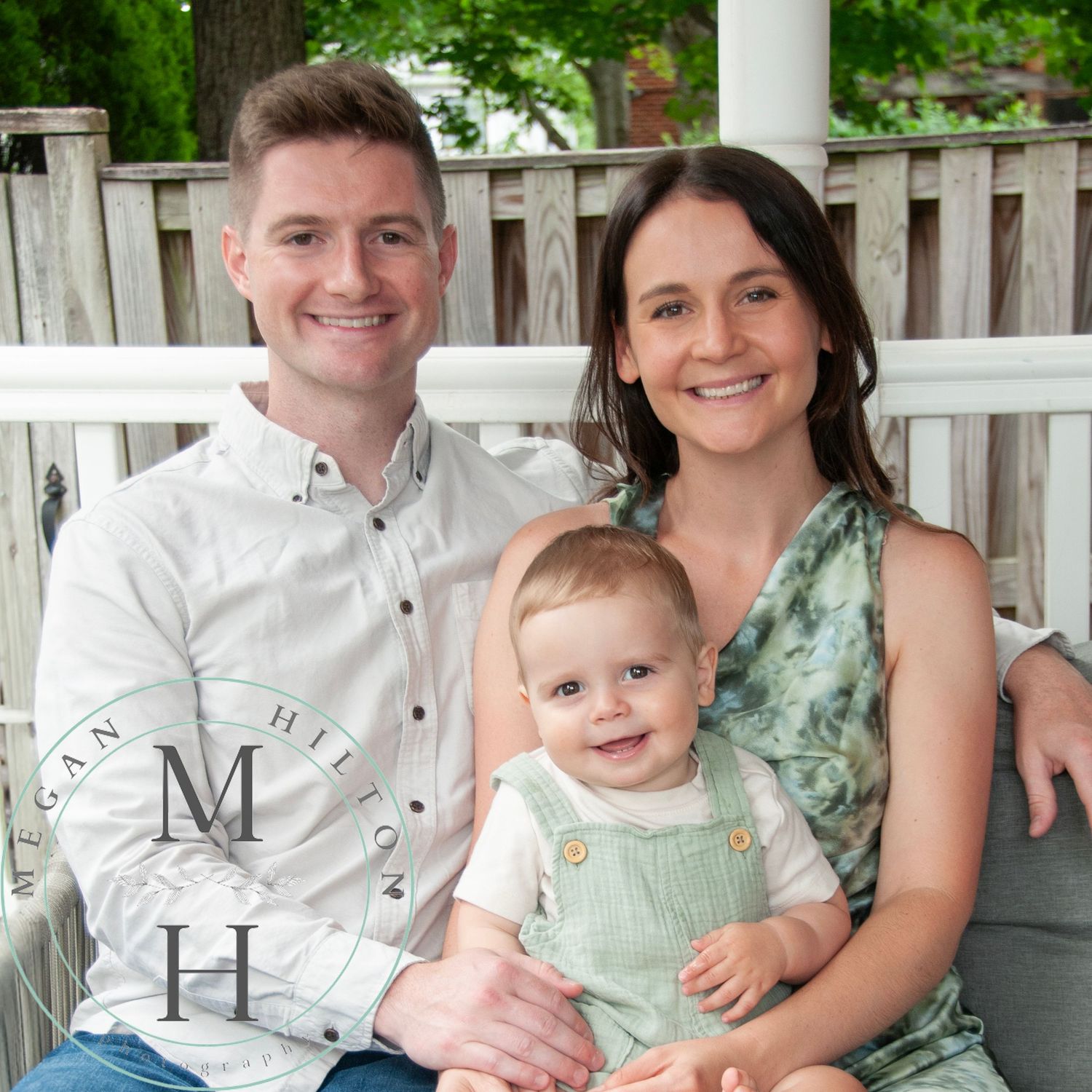 A young family sits together on a porch with white fence and greenery in the background.