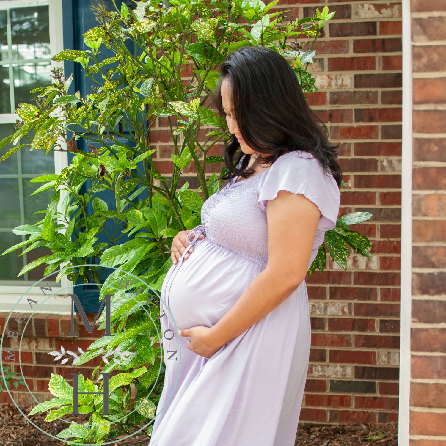 Picture of a pregnant mom near a brick wall and green plants while cradling her pregnant belly.