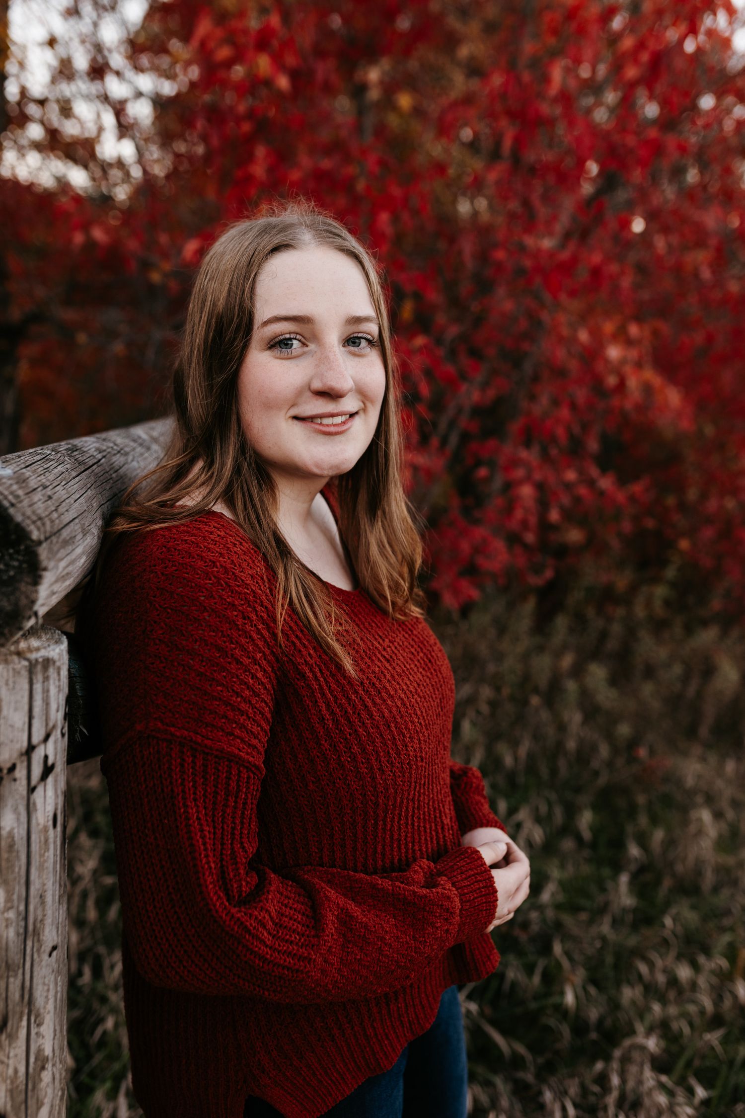 Someone poses outdoors in a burgundy sweater against vibrant red autumn foliage.
