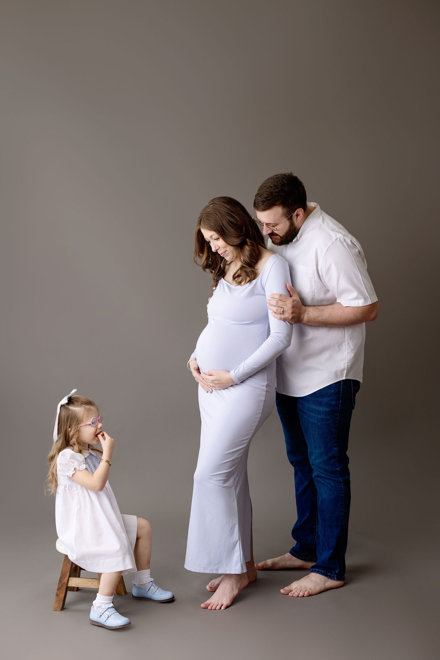 Family maternity photo featuring parents and young daughter in white outfits against neutral gray backdrop in studio setting.