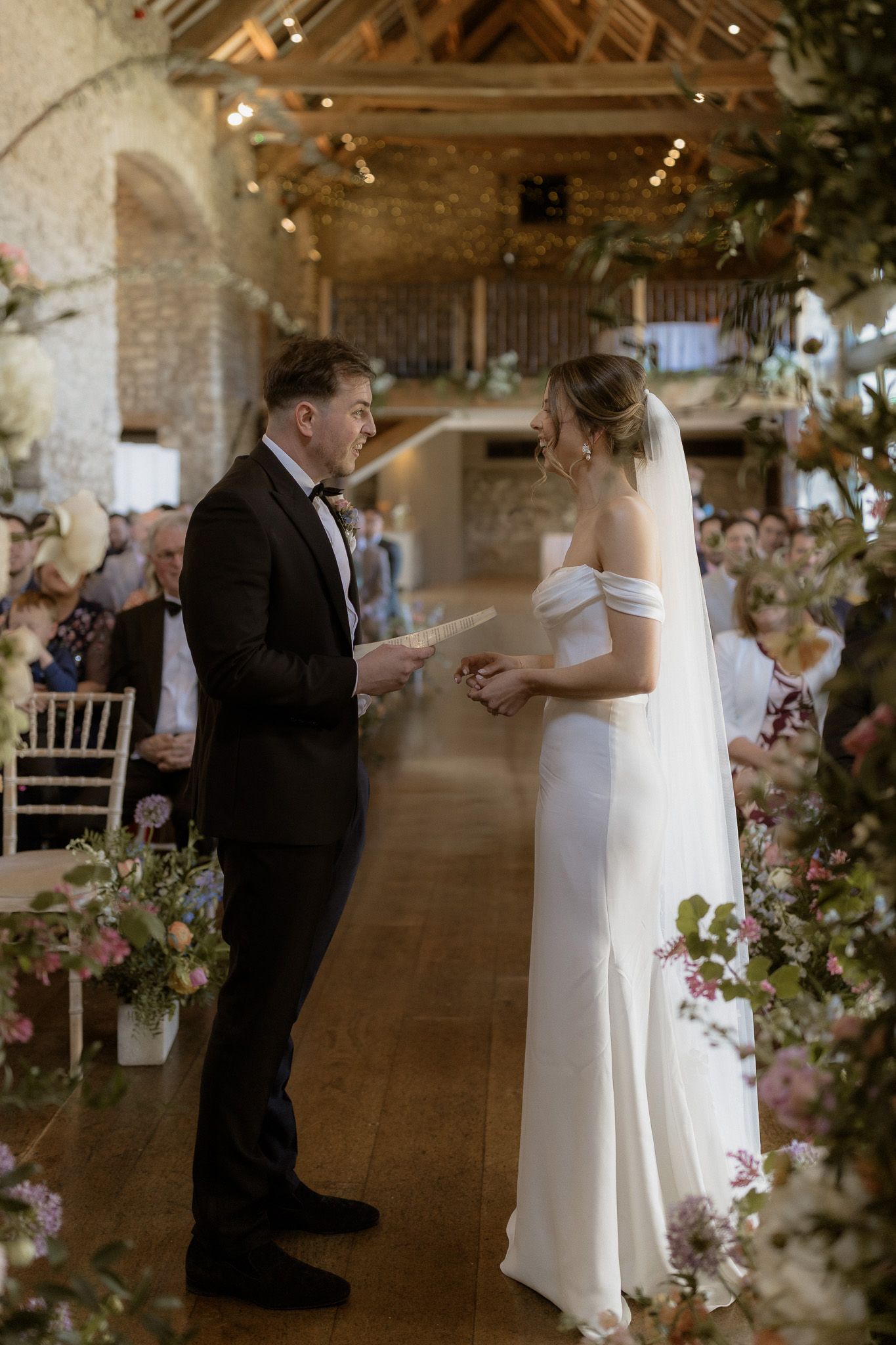 Couple exchanging vows during wedding ceremony in rustic barn venue adorned with floral decorations.