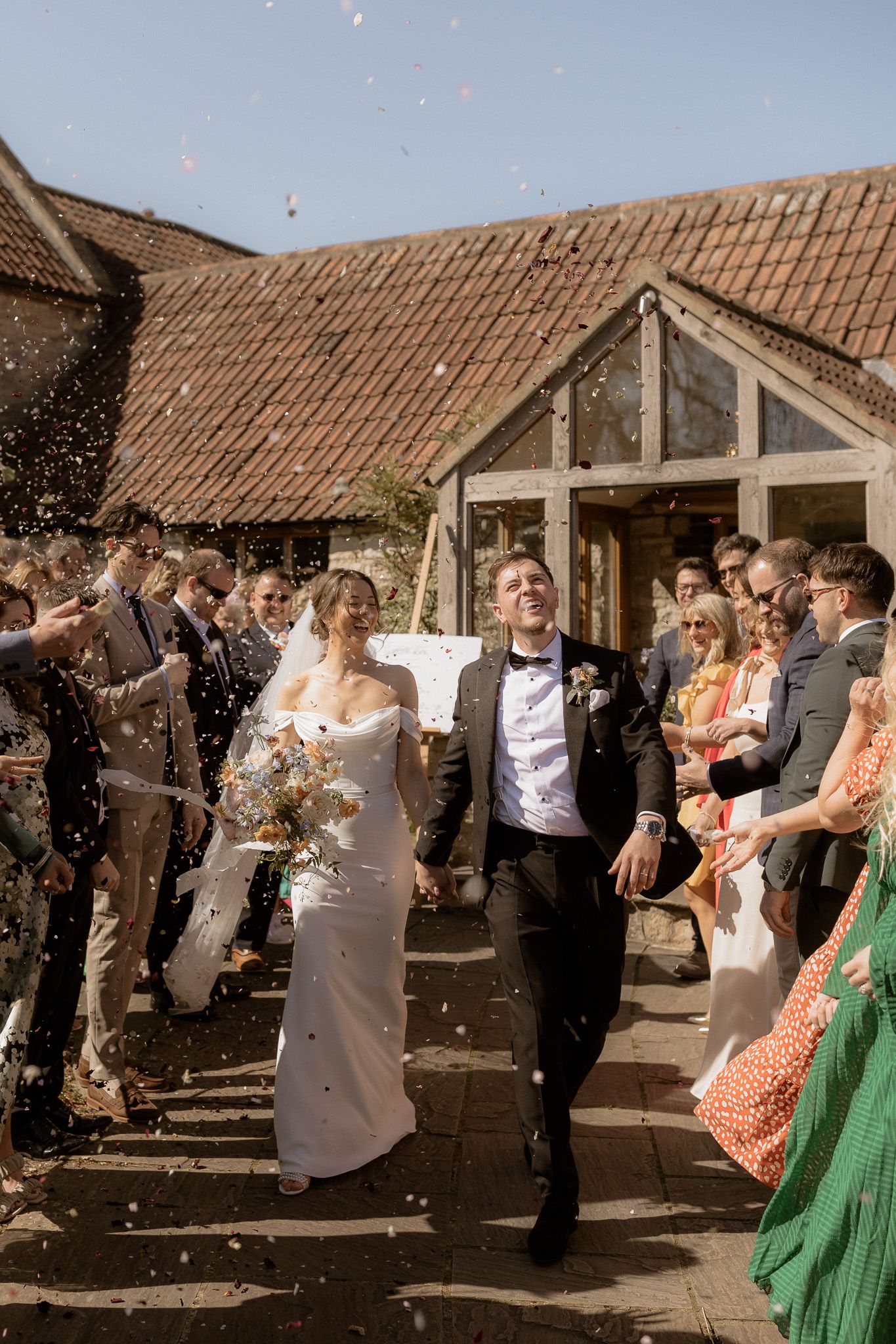 Newlyweds exiting wedding venue while guests shower them with confetti in golden sunlight.